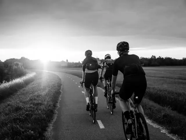 Drie wielrenners van achteren op een fietspad langs akkers bij zonsondergang
